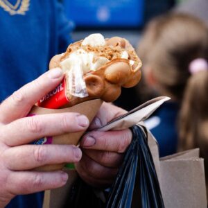 A person holds a bubble waffle topped with cream and a Biscoff biscuit, with a crowd in the background.