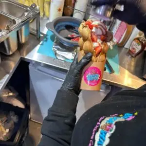 A gloved hand holds a freshly made bubble waffle topped with strawberries and cream inside the dessert bar.
