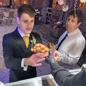 A groom in a black suit receives a bubble waffle at a wedding, with another guest holding one in the background.