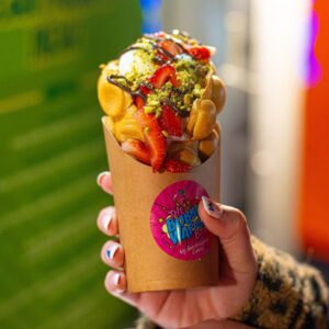A hand holds a bubble waffle topped with strawberries, pistachio, chocolate drizzle, and cream.
