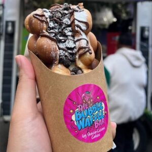A hand holds a bubble waffle topped with crushed cookies, chocolate drizzle, and icing sugar.