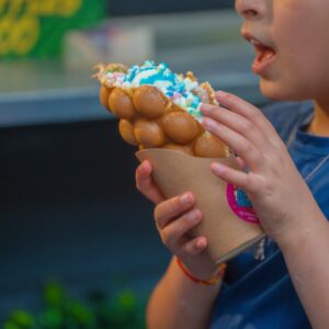 A close-up of a child holding a colourful bubble waffle with blue ice cream and mini marshmallows.
