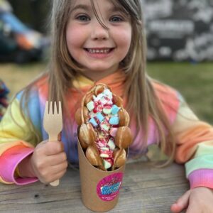 A smiling young girl in a rainbow hoodie holding a colourful bubble waffle topped with sprinkles and marshmallows.