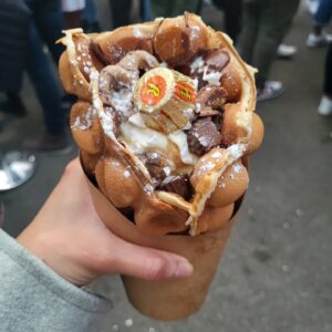 A hand holds a chocolate-loaded bubble waffle topped with Reese’s peanut butter cups and icing sugar.
