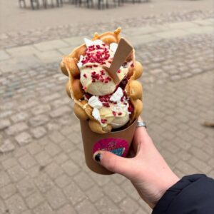 A hand holds a bubble waffle topped with vanilla ice cream, crushed meringue, and freeze-dried raspberries.