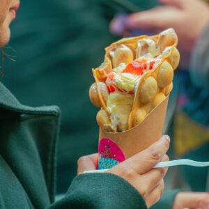 A person holds a bubble waffle filled with cream and strawberries at a food stall.