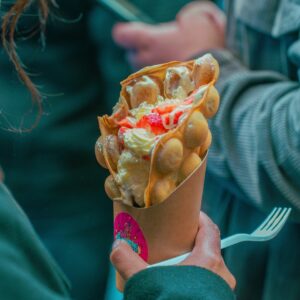 A person holds a bubble waffle filled with cream and strawberries at a food stall.