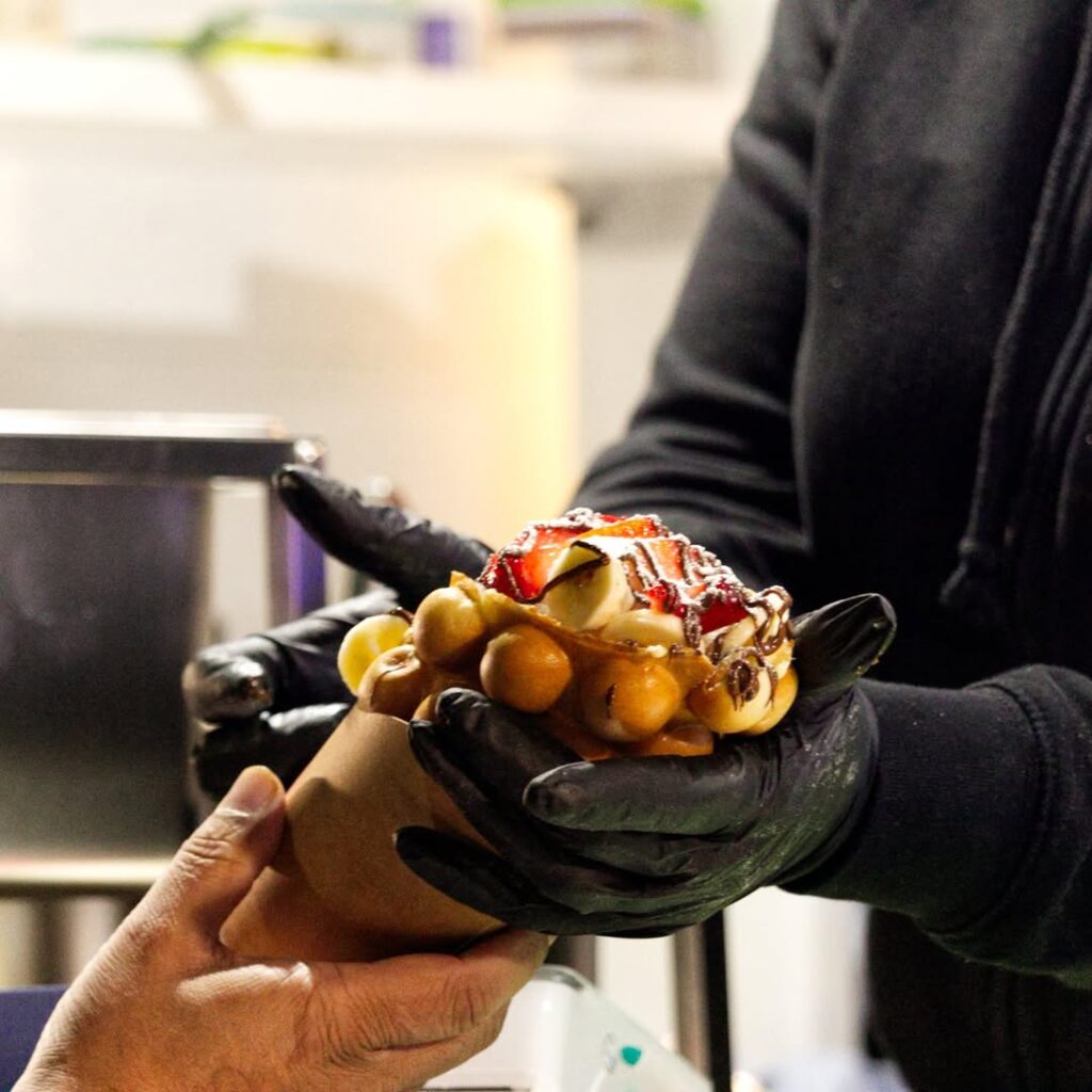 A gloved staff member hands a freshly prepared bubble waffle to a customer.