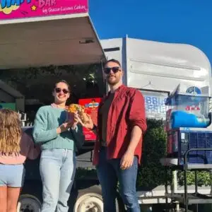 A smiling couple stands in front of The Bubble Waffle Bar holding bubble waffles under bright blue skies.