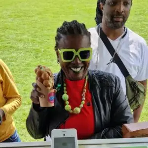 A smiling woman in colourful accessories and green sunglasses holds a bubble waffle at an outdoor event.