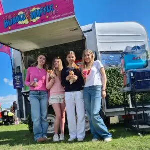 Four young women pose in front of The Bubble Waffle Bar, smiling with waffles and drinks on a sunny day.