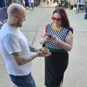 A man holding a bubble waffle smiles at a woman checking her phone at an outdoor food event.