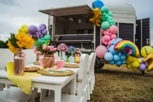 A pastel-decorated horsebox with balloon garlands and unicorn/rainbow details, alongside a long kids’ table set for a party.