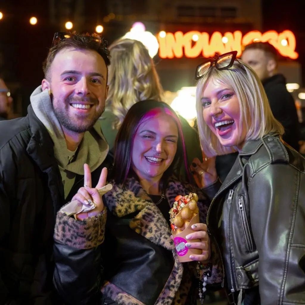 Three smiling friends pose at a night market with a bubble waffle in hand, surrounded by lights and crowd.