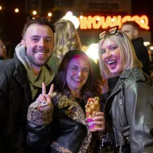 Three smiling friends pose at a night market with a bubble waffle in hand, surrounded by lights and crowd.